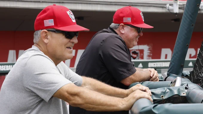 Two men wearing red baseball caps lean on a padded railing, watching a baseball game from the dugout area.