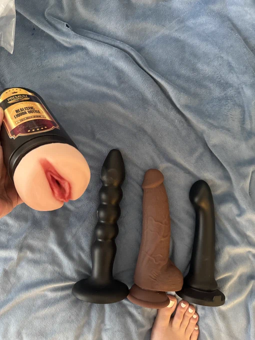 3 adult toys lay on the bed above Fiffany’s foot and in their hand is a pale pocket pussy with pink lips. Their toenails are pained white.