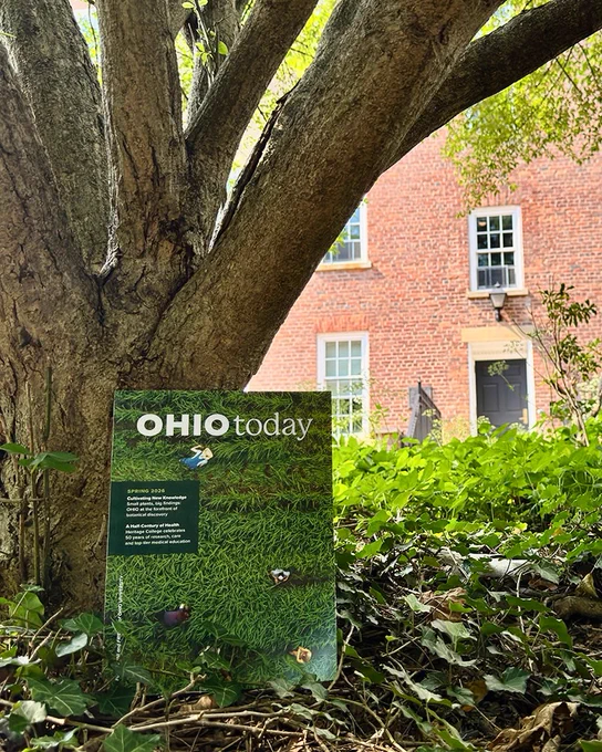 The image shows a copy of "Ohio Today" magazine placed upright against the base of a large tree trunk. The magazine cover features an aerial view of green grass with small people walking. In the background, there is a brick building with white-framed windows and a door, partially obscured by foliage.