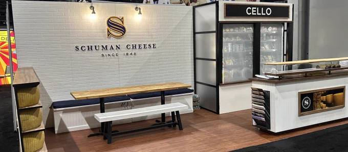 A trade show booth for Schuman Cheese, featuring a display for Cello products. The booth has a rustic and professional atmosphere with wooden tables and cheese wheels.