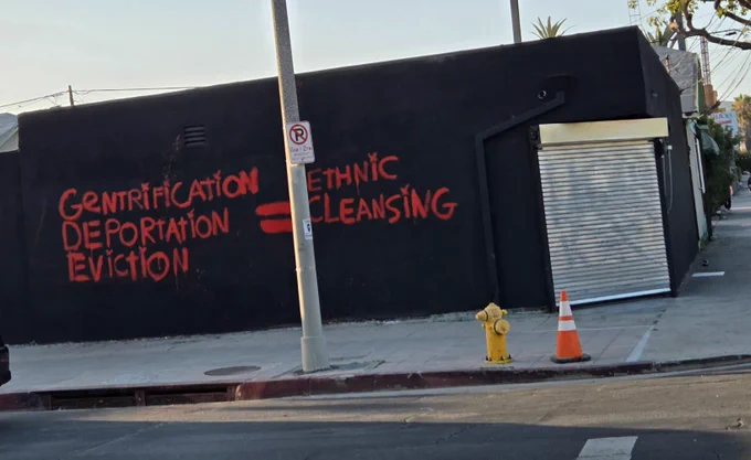 Red spray paint on a black building reading "GENTRIFICATION 
DEPORTATION
EVICTION

=

ETHNIC CLEANSING"
