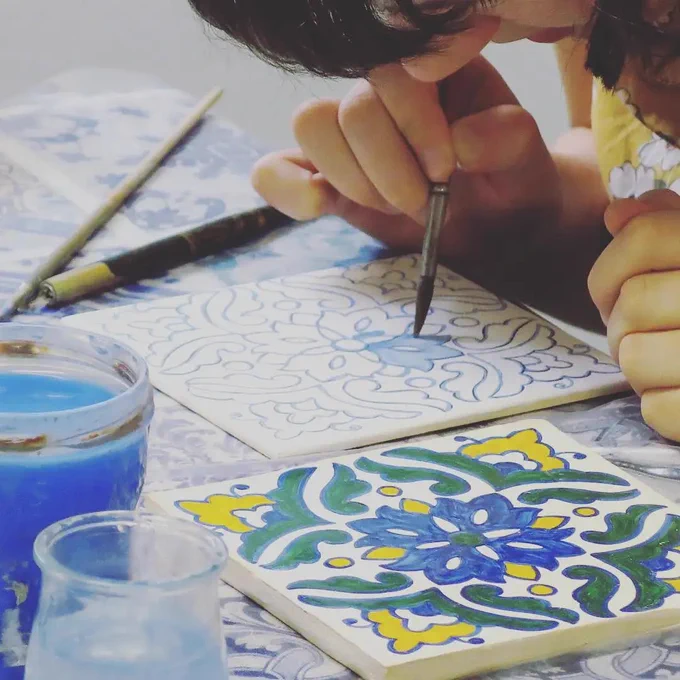A person painting a traditional blue, green and yellow azulejos-style ceramic tile by hand, with paint pots and brushes arranged on a patterned table.