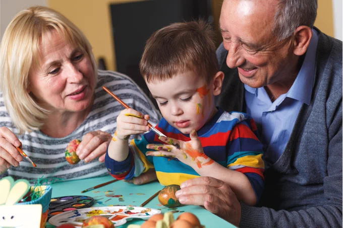 A young boy paints Easter eggs with older couple, with paint on his face and hands.
