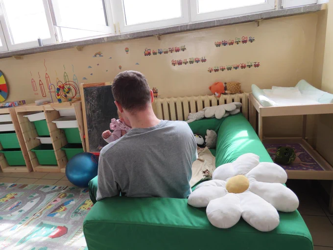 Mężczyzna trzymający w dłoniach dziecko,  w tle ozdoby kącika dziecięcego i zabawki
