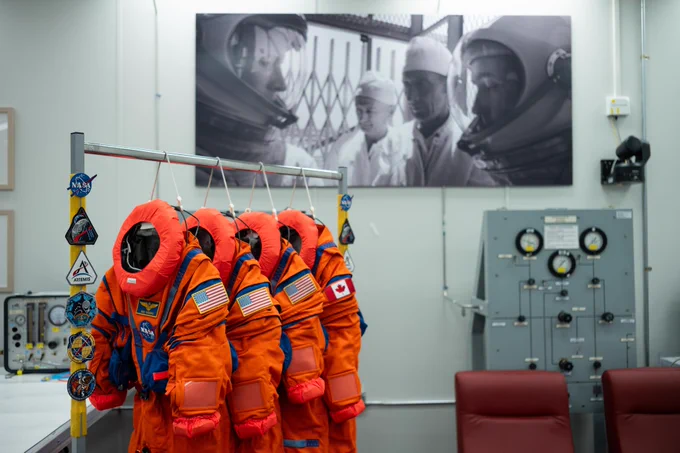 Los cuatro trajes anaranjados del Sistema de Supervivencia de la Tripulación de Orion colgados en la sala de preparación de trajes del Edificio de Operaciones y Revisión Neil A. Armstrong, en el Centro Espacial Kennedy en Florida. Los trajes, que fueron usados por la tripulación de Artemis II, tienen parches de NASA, Artemis y banderas de Estados Unidos y Canadá. Al fondo se observa una fotografía en blanco y negro de personal con equipo de protección, junto con paneles de control y sillas rojas dentro de la sala. 