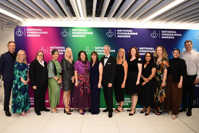 A group of people standing in front of a colorful "National Fundraising Awards" backdrop. They are dressed in formal attire, and the atmosphere appears celebratory. The backdrop features repeated logos of the event.