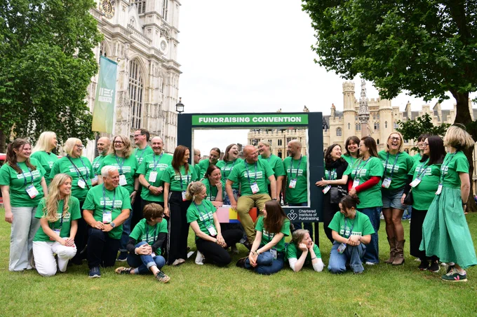 A group of people wearing matching green shirts poses outdoors at a fundraising convention. The setting includes large historic buildings and trees. A sign in the background reads "Fundraising Convention" and features the logo of the Chartered Institute of Fundraising.