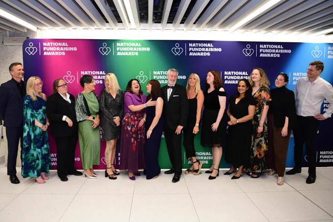 A group of people standing in front of a colorful backdrop that reads "National Fundraising Awards." They are dressed in formal attire, smiling, and interacting with each other. The backdrop features logos and text with a heart design.