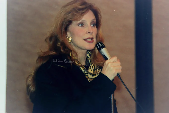 Gates McFadden with her long amazing unicornish long strawberry blonde hair falling on shoulders. She's wearing a big black coat, gold earrings and a yellow/brown/black scarf