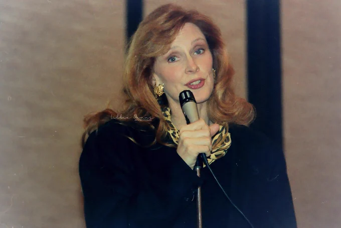 Gates McFadden with her long amazing unicornish long strawberry blonde hair falling on shoulders. She's wearing a big black coat, gold earrings and a yellow/brown/black scarf
