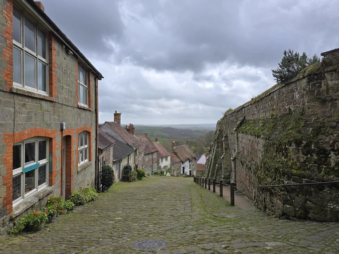 Zu sehen ist eine recht schmale Straße in einem kleinen englischen Dorf. Auf der rechten Seite ist eine Mauer zu sehen, die teilweise mit Moos bewachsen ist. Auf der linken Seite sind kleine, eng zusammenstehende Häuser zu sehen. Die Straße ist abschüssig, der Blick geht nach unten. Es sind keine Autos oder Menschen zu sehen.
Der Himmel ist stark bewölkt.