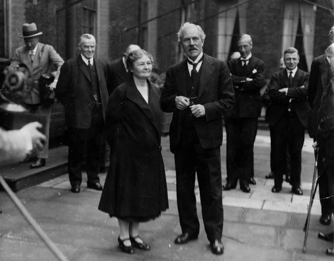 A group of well-dressed individuals stands outside, with one person in the front appearing to speak. They are surrounded by people wearing suits. The setting appears formal and historical.