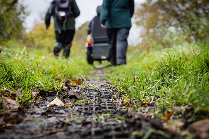 People in outdoor clothing walk along a narrow, muddy path through grass and fallen leaves, carrying fieldwork equipment, including a large case, with one person wearing a backpack. The photo is taken from a low angle with the foreground in sharp focus—showing wet soil, grass, and leaf litter—while the people and background are softly blurred, suggesting a natural riverside or countryside setting.