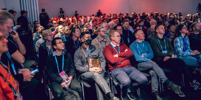 Large audience attentively watching a presentation in a dimly lit conference room with red overhead lighting.