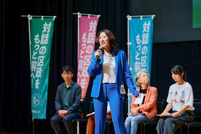 ステージ上で、鮮やかなブルーのスーツを着用した岸本さとこが、マイクを持って力強くスピーチをしている様子。背後には「対話の区政を もっと前へ！」と書かれたピンクや青、緑の応援旗（のぼり）が立ち、その前で他の登壇者が着席して話を聞いている。