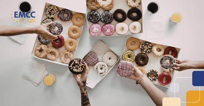 Four hands reaching for assorted donuts in open boxes on a white table with coffee and orange juice cups nearby.