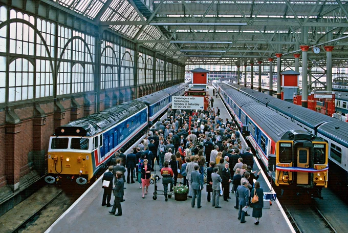 On the left of the picture a red white and blue class 50 locomotive on the right a red white and blue class 455 , the right hand train has a similar coloured ribbon around it's front.
The platform is in-between the trains.
The platform is full of people inspecting the new livery. 
All this takes place under the glass roof of Waterloo station 