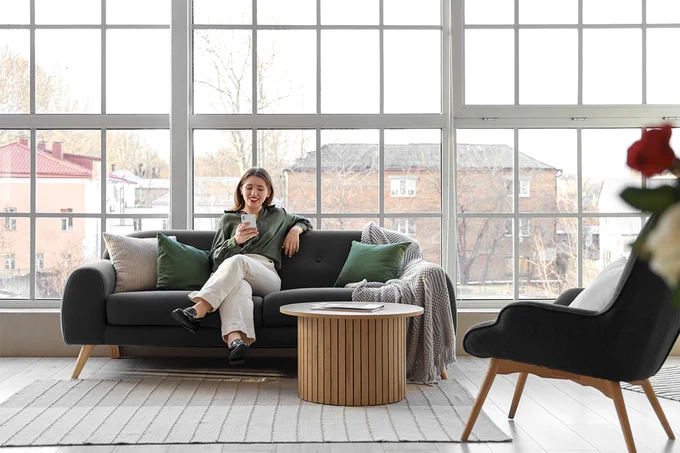 A person is sitting on a gray sofa, holding a smartphone, in a bright room with large windows. A modern coffee table and an armchair are nearby. The view outside shows some buildings and trees.