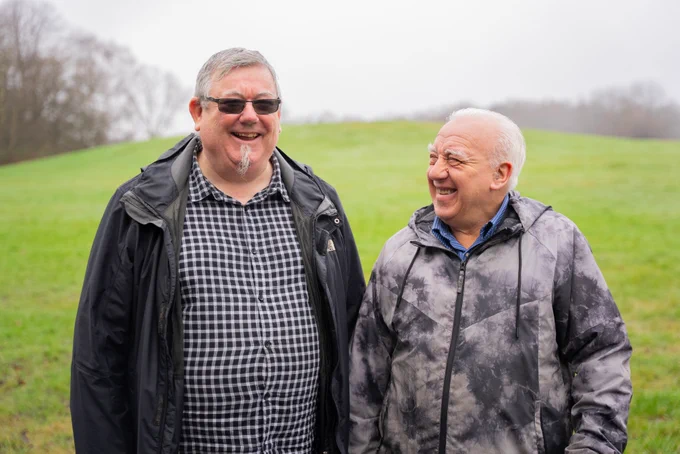 Two smiling men in outdoor jackets stand in a grassy field.