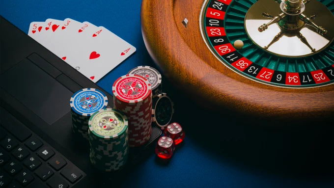 A roulette wheel, playing cards, poker chips, dice and a laptop on a blue table. 