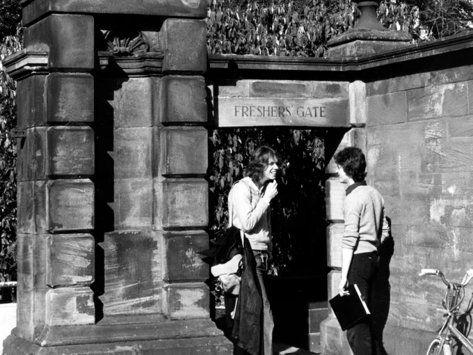 A black-and-white photo shows two individuals standing near a stone structure labeled "Freshers' Gate." One person holds a book while the other appears to be mid-conversation. A bicycle is partially visible nearby. Dense foliage forms the backdrop.