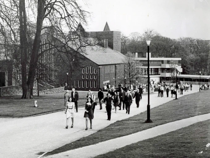 A group of people walking on a paved path surrounded by grass on a university campus. In the background, large brick buildings are visible, with trees lining the area.