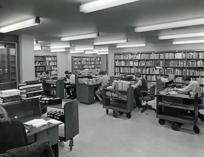 A large library room with several people seated at desks covered with papers and books. Bookshelves filled with books line the walls. The room is well-lit with fluorescent lights on the ceiling.