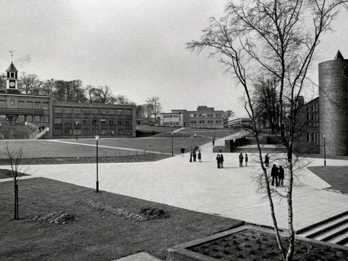A group of people are walking across a large, open courtyard surrounded by modern university buildings. A clock tower is visible on one of the buildings. The scene has a calm, academic atmosphere. Trees are scattered around the area.