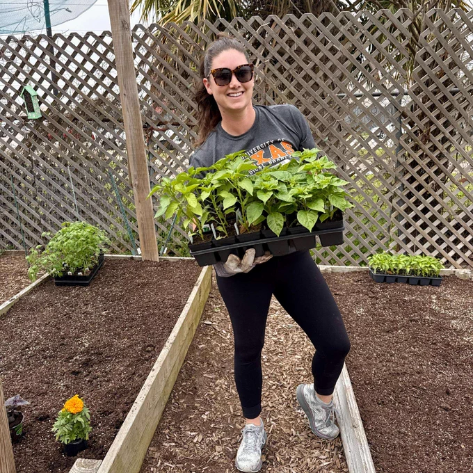 A person is smiling and holding a tray of young plants in a garden area. They are wearing sunglasses and standing on a path covered with wood chips. Raised garden beds with soil and plants are visible, along with a trellis in the background.