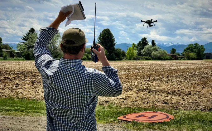 Man in checkered shirt operating a drone with a remote control near a field and a drone landing pad on the ground.