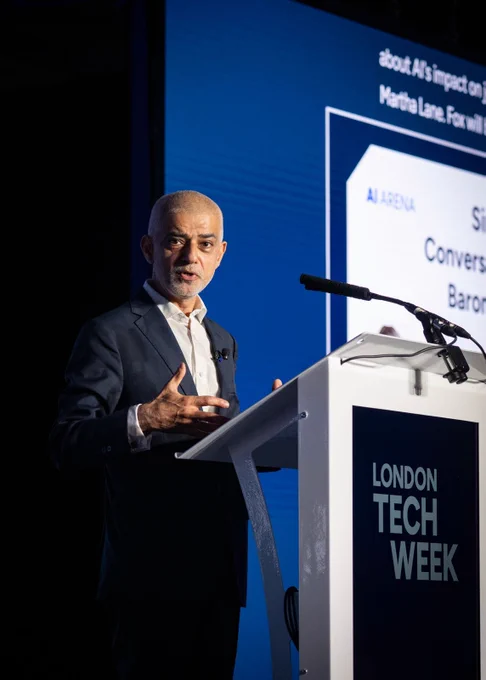 Mayor of London Sadiq Khan in a dark suit stands behind a clear lectern on a stage, speaking into a microphone and gesturing with one hand. A large blue screen fills the background with partially visible presentation text and a slide. The lectern displays the words “LONDON TECH WEEK” in bold white letters.
