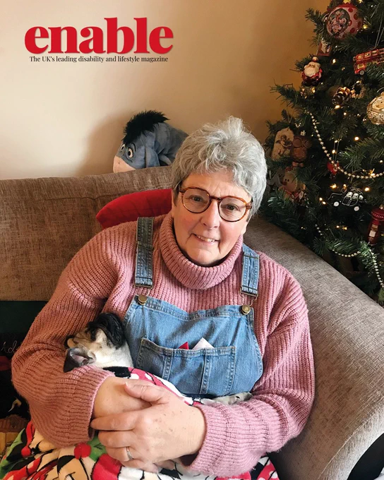An image of Liz and her pug Jarvis. Liz is smiling, has glasses and grey hair, and is wearing a pink knitted turtleneck and denim overalls. She is holding Jarvis and sitting on a sofa with a Christmas tree in the background.
