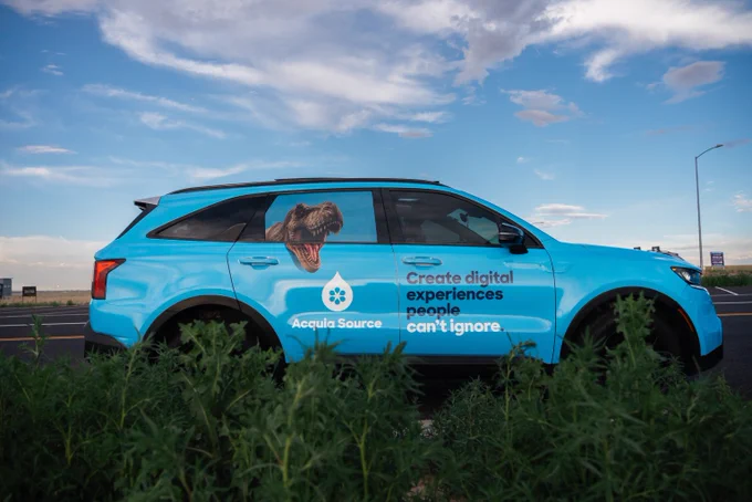 Bright blue SUV with Accula Source branding and a dinosaur graphic parked by the roadside under a partly cloudy sky.