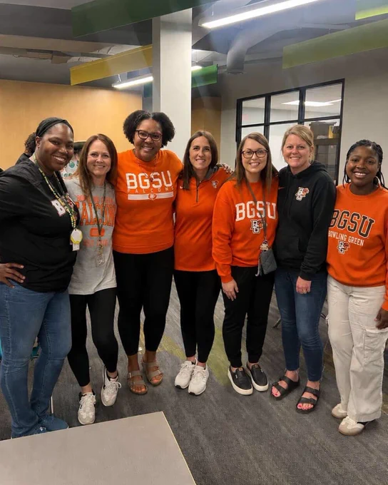 A group of seven people standing together and smiling. Some are wearing orange or black "BGSU" apparel. They appear to be in an indoor setting with a modern design.