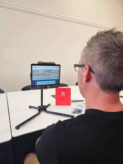 A man viewing a communication device with an eye tracker, mounted on a tripod stand displaying a colourful vocabulary set. The setting includes a white table with a red booklet.