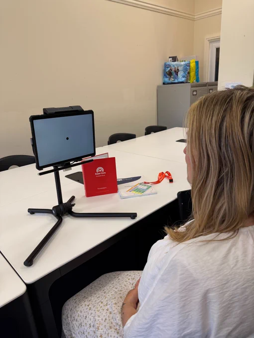 A woman sitting at a table facing a communication device with an eye tracker, mounted on a tripod stand. The woman is practicing screen calibration.
