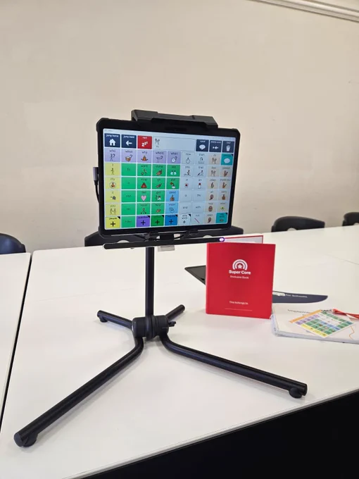A communication device with a grid of colourful icons on a screen, mounted on a black tripod stand placed on a white table. Next to the device are a red booklet labelled "Super Core" and some printed materials.
