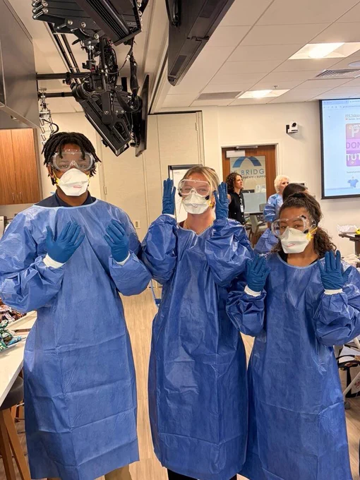 A group of students wearing full protective gear—blue gowns, gloves, masks, and goggles—stand in a clinical training room. Three students pose at the front while others work at tables behind them. A ceiling‑mounted monitor and classroom equipment are visible in the background.