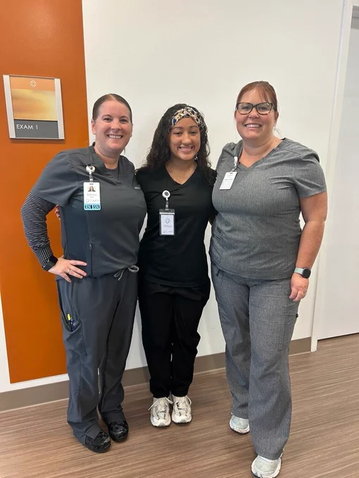 Three individuals in medical scrubs stand side by side in a clinical hallway near a sign labeled “Exam 1.” They smile toward the camera, each wearing ID badges. The setting includes wood‑patterned flooring and a white wall with an orange accent panel.