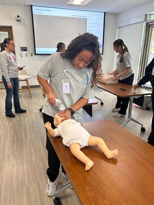 A student practices infant CPR techniques on a baby manikin, holding a resuscitation mask over the manikin’s face. Other participants work with training equipment in the background, and a presentation slide about choking relief is displayed on a screen.