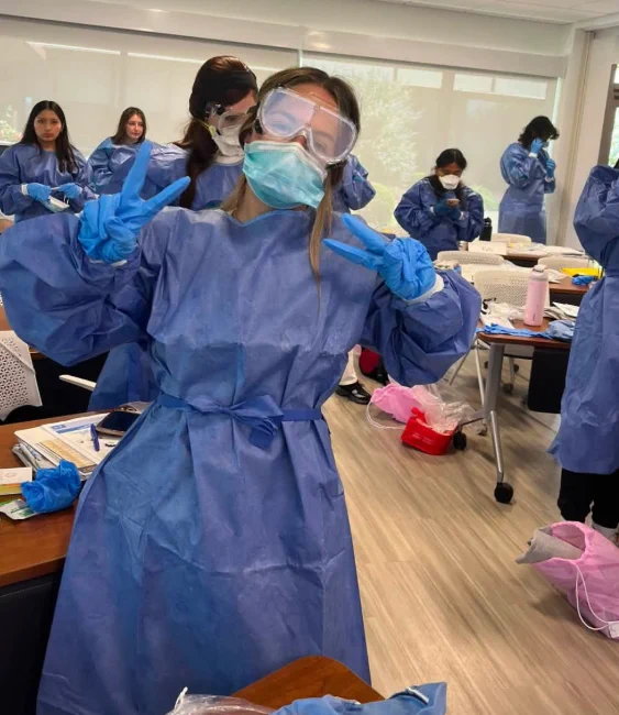 A student in full protective gear—blue gown, gloves, mask, and goggles—stands in a classroom making a peace sign with both hands. Other students in similar gear work at tables covered with papers and supplies.