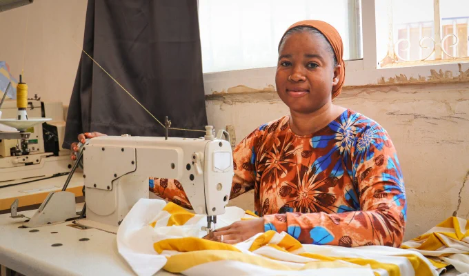A person is seated at a sewing machine, working on a piece of fabric. The room has a window with a partial curtain, and an industrial sewing machine is visible. The person is wearing a colorful floral-patterned top and headwrap.