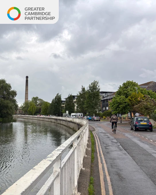 Riverside road beside a curved metal railing with calm water, a cyclist riding along a road with parked vans, trees lining the street, and a tall chimney in the distance under a cloudy sky.
