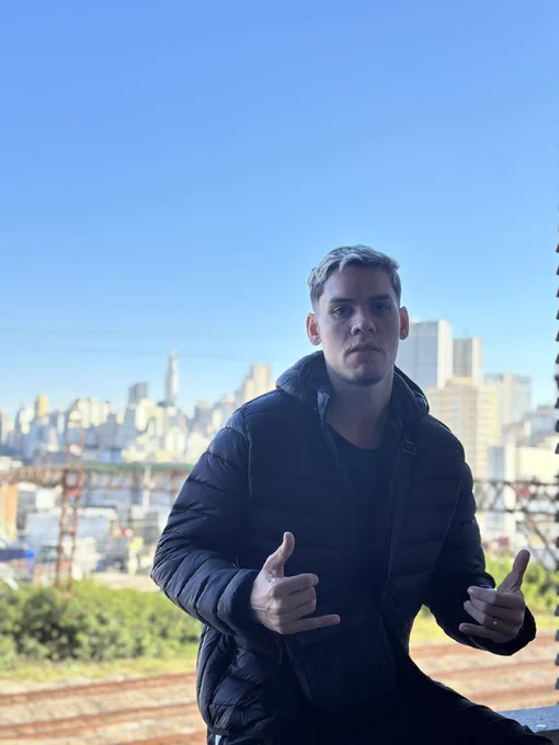 Homem de jaqueta preta sentado em murro por traz cidade de sao paulo e os trilhos dos trens