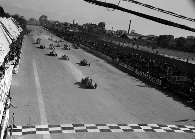 Shot from high up in the pit building, a black and white photo of Alberto Ascari trailing dust or smoke in his Ferrari 375 as he heads for the chequered start/finish line, the rest of the field in a straggly formation behind him. Spectators fill the low grandstands on either side of the track. 1950 Penya Rhin Grand Prix, Michael Tee.