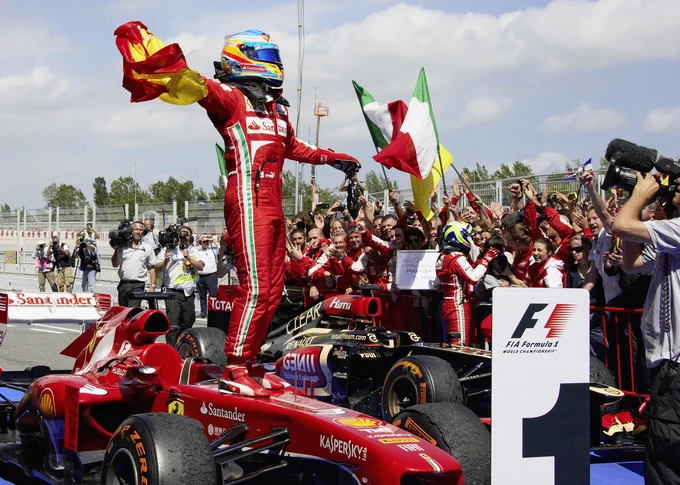 On the centre left of frame, Fernando Alonso stands on his red Ferrari F138 in parc ferme, clutching a Spanish flag in his right hand as he celebrates. He wears a red race suit with an Italian flag stripe down the side, and a blue, yellow and red crash helmet. The Ferrari team stands at the barriers behind, some waving Italian and Ferrari flags. 2013 Spanish Grand Prix, Rainer Schlegelmilch.