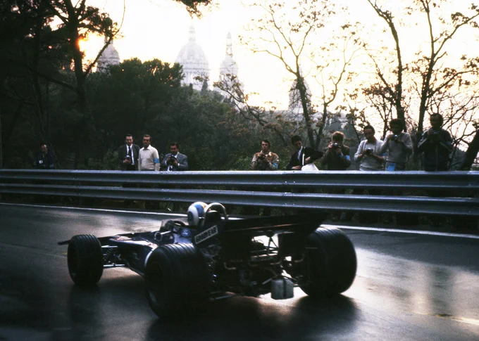 Under a sky fiery from the low evening sun, François Cevert passes from right to left of frame in his dark blue Tyrrell 002, the damp road surface reflecting the car's shape. Ten photographers line the crash barrier in the background, trees behind them. 1971 Spanish Grand Prix, Rainer Schlegelmilch.