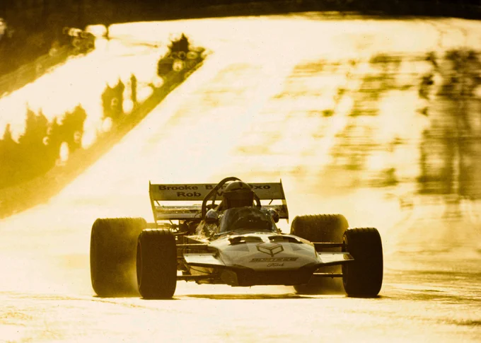 The scene glowing from a golden evening sun reflecting on the damp road surface behind, John Surtees is on track in his Brooke Bond Oxo sponsored, blue and white Surtees TS9. He heads for slightly right of the camera. 1971 Spanish Grand Prix, Rainer Schlegelmilch.