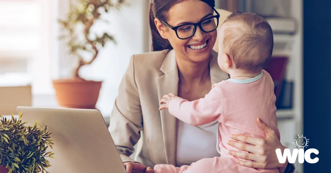 Woman in business attire holding a baby while sitting at a desk with a laptop and plants nearby.