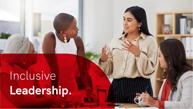 Diverse team of business women talking and brainstorming around a table in an office. In the bottom left corner is a red overlay with white text reading: 'Inclusive Leadership.'